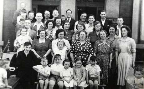 Photograph of a group of twelve men, eleven women, and eleven children posed outside the entrance to a hotel (a sign reading Open To Residents can be seen behind the group) in an unidentified location; the photograph has been identified as portraying a group of holiday-makers, from Shotton, in Blackpool