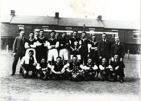 Photograph of eleven men in football strip, posed with eight other men, in front of goal posts, with terraced houses in the background; a man on the front row is holding a football and twelve medals; it is not possible to read the writing on the football, but the photograph is identified as portraying Shotton Imps Football Club