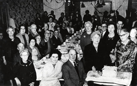 Photograph of approximately fifty elderly people standing behind, and in front of, a long table covered with a cloth and looking at the camera; streamers and paper bells are hanging at the back of the room so the occasion is, no doubt, a Christmas party; at the front of the picture, a woman is about to cut a large iced cake; the photograph has been identified as Old Age Pensioners' Association, Shotton