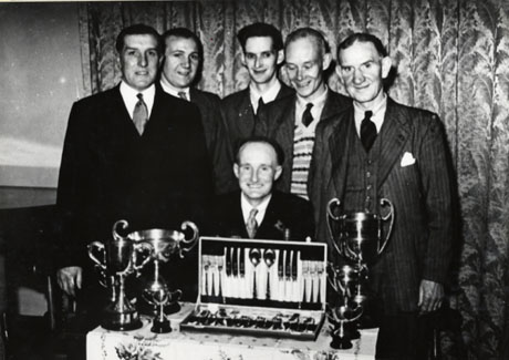 Photograph of six men standing and one man sitting in front of a curtain and behind a table on which trophies stand; all the men are wearing suits and ties; the trophies are four large cups, one small cup and a canteen of cutlery; the photograph has been identified as Homing Pigeons Society Prize Giving