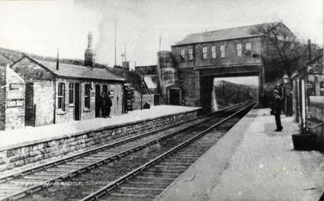 Postcard photograph entitled Shotton Bridge Station, showing the railway line running away from the camera to the right; both platforms and the buildings on each platform can be seen; a bridge consisting of a building with windows can be seen; indistinct figures are on the platforms