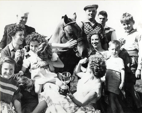 Photograph of a group of four women, five children, three men, surrounding a horse, the head of which, only, can be seen; one woman is holding a large trophy cup and another a small trophy cup; the photograph has been identified as showing Head Horse Keeper, R. Young, with the pony, Tom, at Thornley Carnival