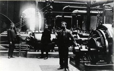 Photograph of three men standing in front of machinery inside a large building; the machinery and building have been identified as the Exhauster House at the Coke Ovens at Shotton; the machinery, which appears to consist of long cylindrical engines, and pipes rising from them, can be seen