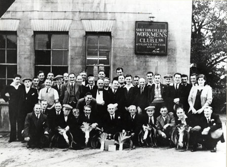 Photograph of a group of forty one men, one boy and one woman, grouped outside a building bearing the sign: Shotton Colliery Workmen's Club, Ltd. Affiliated to Club Union; in front of the group are seven large leeks and a small trophy cup on a box; the group has been identified as Shotton Colliery Palms Leek Club; the woman has been identified as Winnie Henderson