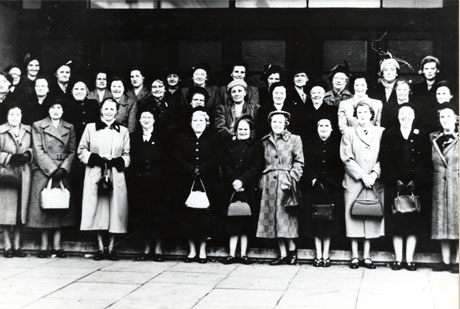 Photograph of thirty six women wearing overcoats and hats and carrying handbags, grouped outside indistinctly seen large glass doors; they have been identified as Shotton Colliery Women's Institute trip to Paton and Baldwin's factory
