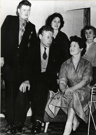 Photograph of two men and three women, members of Shotton Colliery Amateur Dramatic Society; one man, dressed in blazer and tie, is standing on the left; the second man, dressed in jacket, cardigan and tie, is sitting on the left arm of the chair in which one of the women, who is dressed in a smart frock, is sitting; behind the seated man and woman the other two women are standing; the man on the extreme left has been identified as John Winning, and the woman on the far right has been identified as Mrs. Joan Harker