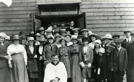 Photograph of approximately forty people standing in the doorway of a wooden building, behind a clergyman in a surplice; the occasion has been identified as The Opening of Shotton Colliery Roman Catholic Church
