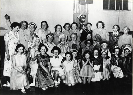 Photograph of sixteen women, ten girls and one boy, posed in a room; a number of the people are in costume and the rest in smart frocks; a woman on the back row has a papier mache dummy, with the appearance of a Black nad White Minstrel, behind her; Mrs. Winnings is the third from the right on the back row; the group has been identified as Shotton British Legion Concert Party Comic Pantomime
