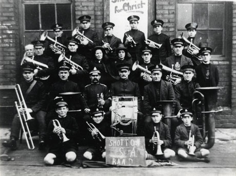 Photograph of seventeen men, five boys and two women in Salvation Army uniform, posed outside a brick building with two windows; eighteen of the group are holding musical instruments; in front of the group is a notice reading: Shotton S. A. Band