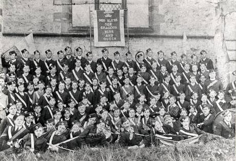 Photograph of approximately one hundred and eight boys in the uniform of the Boys' Brigade, posed against the wall of a church; five men, including a clergyman, are to be seen in uniform to the right and front of the group; they are members of the Shotton Colliery and Southwick Church Lads' Brigade; behind the group is a banner reading: God Bless Our Gracious King And Queen