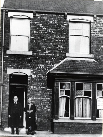 Photograph of an elderly man and woman standing on the pavement in front of the door to a terraced house; the house has two windows on the first floor and a bay window with coloured glass in it on the ground floor; the man and woman have been identified as Mr. and Mrs. Henderson and the house as being situated at Miners' Villas, George Street, Shotton Colliery