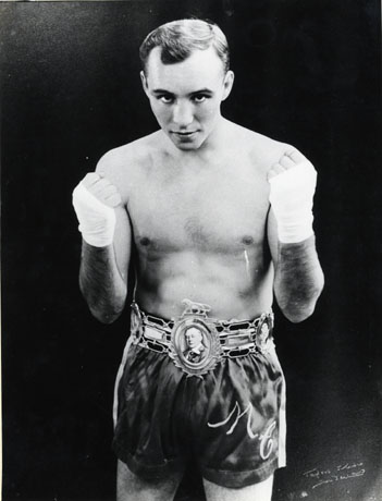 Photograph of a young man stripped to the waist wearing boxing shorts with the initials M. C. on them and with protective bandages on his hands, which he is holding clenched at shoulder height; he is wearing a belt, presumably a trophy, with cameos and an heraldic beast on it, presumably the British Lightweight Trophy ; the young man has been identified as Maurice Cullen, British Champion Boxer; he was British Lightweight Champion five times until he was defeated by Ken Buchanan in 1968; the photograph has the signature of a photographer in Sunderland