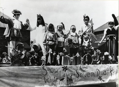 Photograph of ten children in fancy dress standing on a float with the words Snow White and the Seven Dwarfs, written on a cloth hanging down its side; two children aged approximately twelve years are dressed as the Prince and Snow White and six children aged approximately seven years are dressed as the dwarves, and two more children aged approximately twelve years are dressed as a woodman and a young woman; the float has been identified as the Women's Institute Float at Shotton Carnival