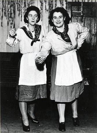 Photograph of two women dressed in skirts, aprons, blouses, and kerchiefs with theatrical scenery behind them; they have been identified as being part of Mrs. Winning's British Legion Concert Party, Shotton and as playing the parts of Irish Colleens; the woman on the left has been identified as Mrs. Short and the one on the right as Mrs. Scobie