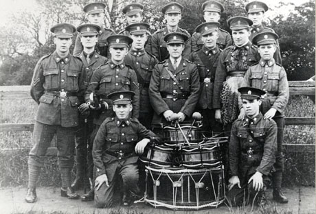 Photograph of eleven young men and three boys in uniform with an officer, posed in the open air in front of a fence and fields; in front of the group is a pile of drums; the group has been identified as Shotton Church Lads' Brigade Band at Shotton Carnival Parade; the man in the middle of the group has been identified as Mr. Scott