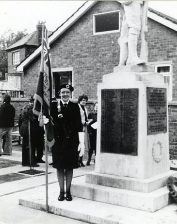 Photograph of the side of the war memorial showing the lists of names and part of the statue of a soldier on the top; the names cannot be read; on the bottom step of the memorial a woman in a uniform holding a flag can be seen; she has been identified as Mrs. Anne Appleby, British Legion Standard Bearer; behind the memorial five figures can be seen and behind them a wall and the side of a house