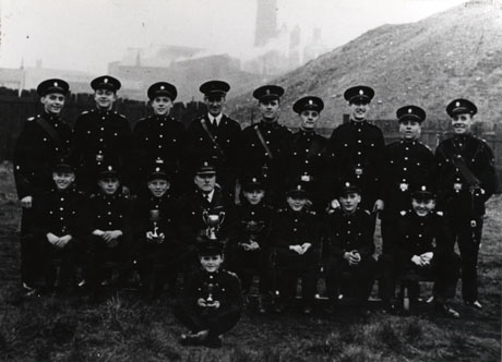 Photograph of fourteen boys and three men in the uniform of the Shotton Colliery Church Lads' Brigade, posed in front of the pit heap with indistinct colliery buildings further in the distance; a man on the front row and three boys are holding trophies, described as Battalion and Camp Trophies