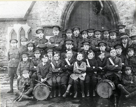 Photograph of thirty one young men and boys in uniform posed against the doorway of a church with part of the exterior wall of the nave in the distance; there are two drums in front of the group, which is described as Shotton Colliery Company Church Lads' Brigade