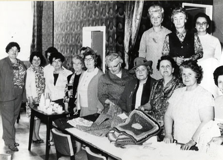Photograph showing two tables lined up at an angle to the camera; on the table nearer the camera are knitted and crocheted garments which are being examined by women standing behind the table; in all there are twelve women standing behind the table and that adjacent, on which tea cups can be seen; three other women are standing on a stage behind the women examining the handiwork; the occasion has been identified as Shotton British Legion Women's Section Arts and Crafts Exhibition, which was opened by Mrs. Davison identified as the woman wearing a hat