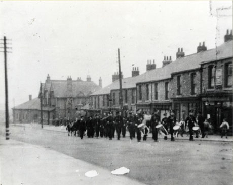 Photograph showing one side of a road, identified as Front Street, running downhill away from the camera; a row of shops can be seen and, at the end of that, an elaborate building identified as The Albert Inn and, next to that, a low building identified as the Colliery Offices; an indistinct parade of people in uniform, identified as a Church Lads' Brigade Whit Sunday Parade, 28 May 1939 is moving up the road