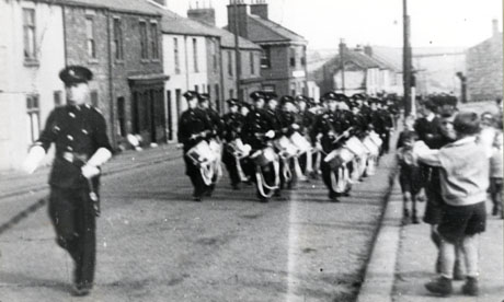 Photograph showing one side of a road, identified as Front Street, Shotton Colliery, running away from the camera to the right; a parade identified as the Church Lads' and Church Girls' Brigade is walking up the road towards the camera; children can be seen on the pavement watching the parade, which is celebrating Wings For Victory in 1943; on the far side of the road, a large building half way down has been identified as The Old Red Brick Pub