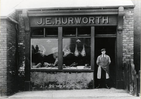 Photograph showing the window of a shop with the name, J. E. Hurworth, above it and large joints of meat in the window; a man wearing a striped apron is standing in the doorway of the shop, which has been identified as being on Potto Street, Shotton Colliery