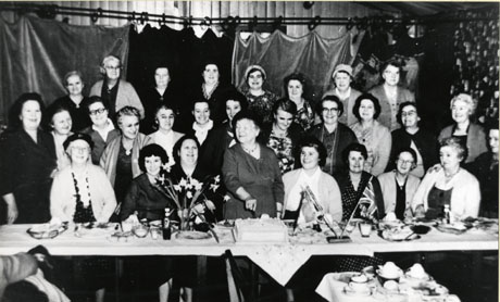Photograph showing a table running across the photograph with eight women sitting behind it and twenty more standing behind them looking at the camera; small flags, a vase of flowers, and the remains of a meal can be seen on the table; in the middle of the group immediately behind the table, a woman is standing up and cutting a large iced cake; the occasion has been identified as Shotton British Legion Women's Section Annual Birthday Party; the woman cutting the cake has been identified as Mrs. Jackson; the woman fifth from the right on the front row as Mrs. Hardy; the woman at the extreme right on the front row as Mrs. Worrall