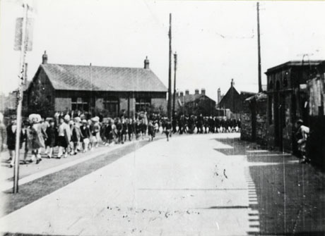 Photograph showing a parade of children walking towards the camera on the left of the picture, passing a single-storey building, with other buildings very indistinct in the distance; the participants in the parade are also indistinct; the building has been identified as The Officials' Club, Shotton Colliery and the parade as Church Lads' Brigade, Whitsunday Parade, 5 June 1938