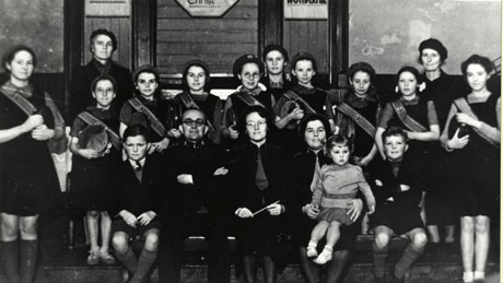 Photograph of fifteen women and girls, with one man and two boys, grouped against a wall inside a building; most of the girls are wearing sashes and holding tambourines; a woman sitting on the front row is holding a small girl; the group has been identified as Shotton Salvation Army-Singing Company