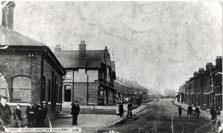 Postcard photograph entitled Front Street, Shotton Colliery. 2128, showing a wide road running away from the camera lined either side with terraced houses and shop fronts; on the left nearest the camera is the side of a single-storey building, identified as the Colliery Offices; approximately thirteen unidentifiable figures can be seen in the foreground