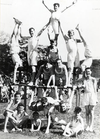 Photograph of sixteen young men and boys in singlets and shorts posing in gymnastic attitudes on a bar set up in a field; crowds of people can be seen indistinctly behind them; they have been identified as Shotton Church Lads' Brigade Gymnastics Display and Road Safety Campaign at Crimdon Park, 1956