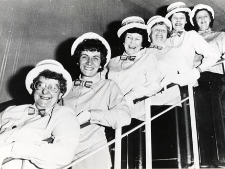 Photograph of six middle-aged women standing on a flight of stairs leaning on the bannisters and posing for the camera; each woman is wearing a shirt, bow tie, and boater; they have been identified as members of Mrs. Winning's Concert Party, Shotton Colliery