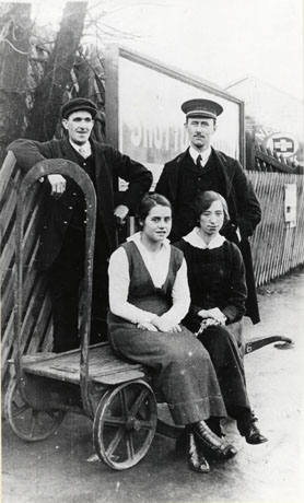 Photograph of two women sitting on a luggage trolley with a man in a railway uniform with a peaked cap, the station master, Mr. Howe, and another man in railway uniform, possibly a porter, standing behind them; a fence and a sign reading, Shotton, can be seen behind the group