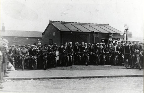 Photograph of approximately fifteen motor cyclists with crowds of people behind them and people on the left of the photograph; behind the motor cyclists and the crowd is a one-storey wood or corrugated iron building with the name Pratt on its side; the building appears to sell petrol, as an early petrol pump can be seen; the building has been identified as a shop, which later became Witton's shop; the motor cyclists have been identified as members of Shotton Colliery Motor Cycle Club