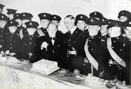 Photograph of sixteen young men in uniform standing behind a long narrow table covered in a cloth and plates of food; in the middle of the picture the senior officer and a man in a dinner jacket are standing near an iced cake which the man in the dinner jacket is about to cut; the event has been identified as the Shotton Church Lads' Brigade Fiftieth Jubilee Party and the man about to cut the cake as Major J. Scott