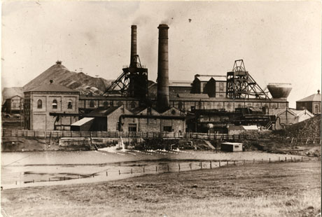 Photograph of the buildings of Shotton Colliery showing the winding house, the winding gear, two chimneys, a pond or reservoir in the foreground and a pit heap in the background
