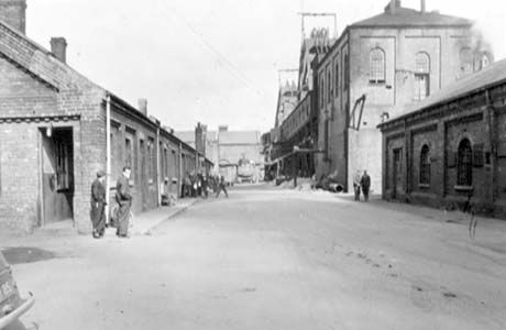 Photograph showing a road running away from the camera between colliery buildings of brick with men standing in front of the building on the left; a building is across the end of the road; the building on the right has winding gear or a similar piece of equipment on it; the colliery has been identified as Shotton Colliery