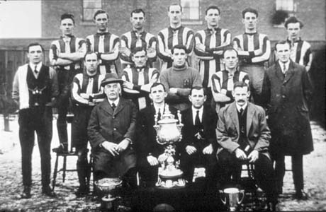 Photograph of eleven men in football strip posed with six other men with houses in the background; in front of them are three trophy cups; they have been identified as Shotton Football Team