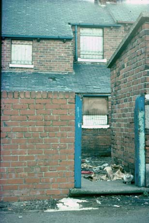 Photograph of a back wall, through the gateway of which, rubbish can be seen on the floor of the yard; the back of a terraced house with boarded up windows and tiles missing from a roof can also be seen; the photograph has been described as View of Back Street, Shotton