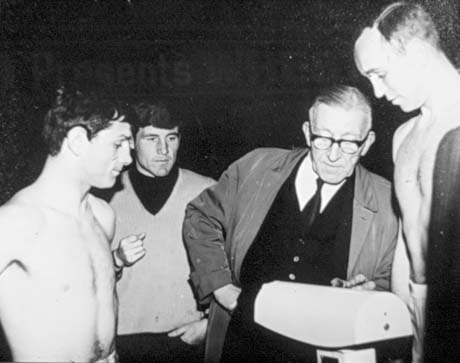 Photograph showing a young man stripped to the waist standing on scales at the right of the picture; a middle-aged man in a raincoat is looking at the scales; a young man in a jumper is also looking at the scales over his shoulder; another young man stripped to the waist is standing at the left of the picture; the picture has been identified as the Weighing In of Maurice Cullen, Shotton