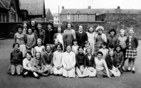 Photograph of thirty one girls, aged approximately between ten and thirteen years, posed in a yard with a wall behind and the front of, possibly a school beyond the wall on the left; houses can be seen in the centre beyond the wall; they have been described as Schoolchildren in Shotton