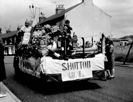 Photograph of the rear of a lorry with the words Shotton Women's Institute on its back; four people can be seen on its back dressed as Red Indians, with a canoe and totem pole; indistinct women are standing on the far side of the float; houses on the far side of the street, along which the float is proceeding, can be seen