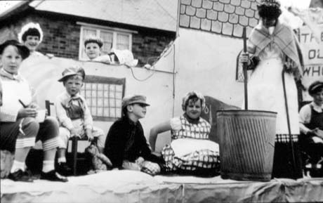 Photograph showing seven children sitting with the wall of a cardboard house with a window and tile roof behind them; a woman, wearing an apron and shawl, is standing with a poss tub; the photograph has been described as Women's Institute Float - The Old Woman Who Lived in A Shoe - Shotton