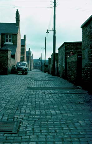 Photograph of a back street running between the back walls of yards of houses and the side ends of houses; the surface of the street is cobbled; a car is parked half way down the street, which has been identified as being in Shotton