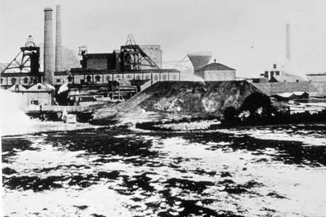Photograph taken from across what appears to be grass covered with snow, of the buildings of a colliery; the winding gear, three chimneys, and a spoil heap can be seen; it has been identified as Shotton Colliery
