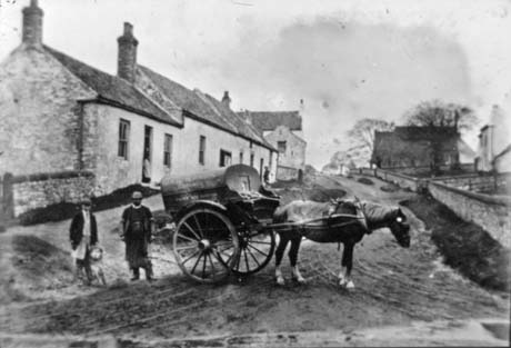 Photograph of a horse harnessed to a cart with a covered container on it; two men are standing near the back of the car; behind them is a row of three cottages; with another building behind; a large building and the walls of gardens can be seen on the right of the picture; the cart has been identified as belonging to Jessie Hutchinson, butcher, who is one of the men standing near the cart, presumably the man on the left with the dog and what appears to be an apron under his jacket