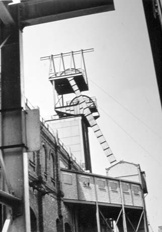 Photograph taken from below of the side of a brick building with an oblong metal superstructure with two wheels above it; it has been described as Skip Winding, Shotton