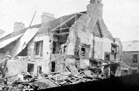Photograph showing the end of a terrace of houses with the roof and wall of the first floor, removed from the end house and the fire places exposed on the end of the building; rubble is lying in quantities on the ground near the houses; the photograph has been described as Demolition of Houses, Shotton; a man can be seen at the left of the photograph