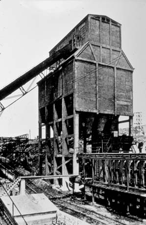 Photograph showing the side of a building on stilts above what have been identified as coke ovens; a railway line runs in front of them; they have been identified as being in Shotton