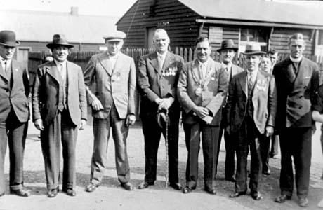 Photograph showing eight men standing in a two with two boys peering over the shoulder of one of them; behind them are a fence with two huts beyond; all the men are wearing suits and ties; two men are wearing military medals and one man is wearing a chain of office; one man is a clergyman in a dog collar; it is possible that the occasion for the gathering, which has been identified as taking place in Shotton, is Armistice Day